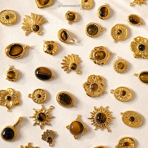 Peut inclure: Une collection de breloques en métal doré incrustées de pierres précieuses œil-de-tigre de formes variées, notamment des rayons de soleil, des fleurs, des ovales et des cercles. Chaque breloque possède une petite boucle, avec le texte @SantomaroDesign.