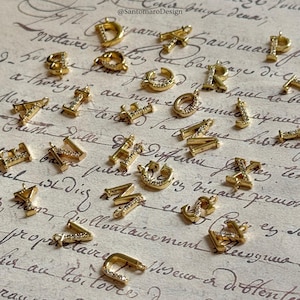 Peut inclure: Charm en forme d'alphabet doré avec de petites pierres transparentes, disposés sur un fond de papier vintage. Les breloques sont en forme de lettres variées, dont les lettres D, A, R et J. Le fond est orné d'une écriture manuscrite.