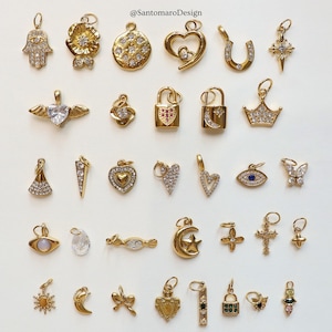 May include: A collection of gold-colored charms with various designs, including hearts, stars, crosses, and a Hamsa hand. Some charms are embellished with clear gemstones. The image includes the text "@SantomaroDesign".
