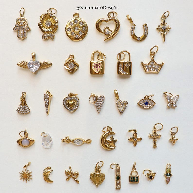 May include: A collection of gold-colored charms with various designs, including hearts, stars, crosses, and a Hamsa hand. Some charms are embellished with clear gemstones. The image includes the text "@SantomaroDesign".