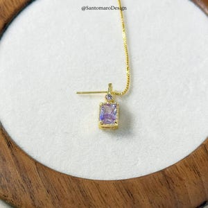May include: A gold-colored earring with a rectangular, light purple gemstone. The earring has a small, round purple stone above the rectangular stone and is suspended from a delicate gold chain. The earring is set against a white background.