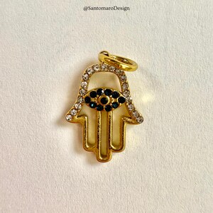 May include: Gold-coloured Hamsa hand pendant with clear and dark blue crystals. The pendant has a detailed design and a loop for a chain. The text "@SantamaroDesign" is in the top left corner.