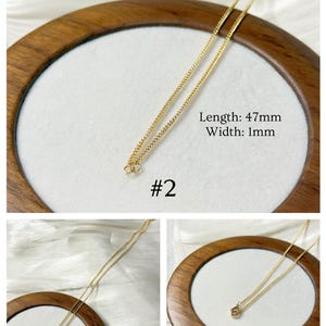 Op de afbeelding: Een delicate goudkleurige ketting. De ketting is 47 mm lang en 1 mm breed. De tekst "Lengte: 47 mm Breedte: 1 mm" en "#2" is inbegrepen. De ketting wordt gepresenteerd op een wit oppervlak in een houten frame.
