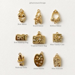 May include: A selection of gold-coloured charms, featuring bikini, makeup bag, and travel bag designs. The charms are intricately detailed with embellishments and are displayed on a light background. The text "@SantomaroDesign" is at the top.