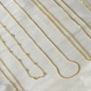 Op de afbeelding: Een verzameling delicate gouden kettingen, gepresenteerd op een witte ondergrond. De kettingen hebben verschillende kettingstijlen, waaronder kralen, slangen en klassieke schakelontwerpen. De afbeelding bevat de tekst "@SantoromaroDesign".