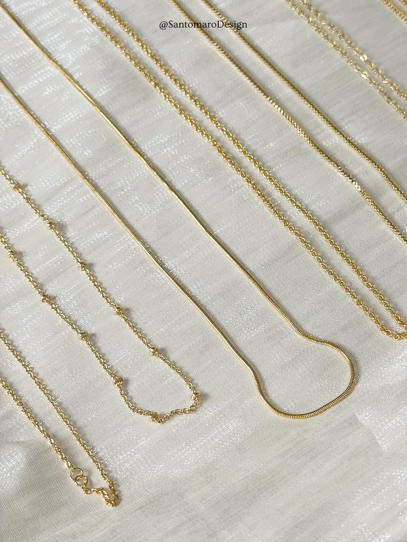 Op de afbeelding: Een verzameling delicate gouden kettingen, gepresenteerd op een witte ondergrond. De kettingen hebben verschillende kettingstijlen, waaronder kralen, slangen en klassieke schakelontwerpen. De afbeelding bevat de tekst "@SantoromaroDesign".