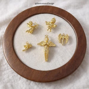 May include: A collection of gold-colored angel charms in various poses, including one with a bow and arrow. The charms are displayed on a white surface within a round, wooden frame. The image includes the text "@SantamaroDesign".