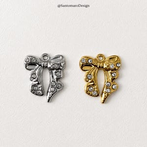 Op de afbeelding: Twee strikvormige bedels, één zilverkleurig en één goudkleurig, elk versierd met heldere strass-steentjes. De bedels hebben een lus aan de bovenkant om op te hangen. De achtergrond is neutraal. De tekst "@SantomaroDesign" staat linksboven.