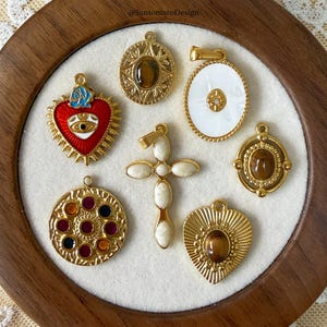 Può includere: Una collezione di ciondoli dorati con vari design. Include un cuore rosso smaltato, una croce con dettagli in pietra bianca e ciondoli con centri in pietra marrone ovali e rotondi. I ciondoli sono esposti su una superficie bianca.