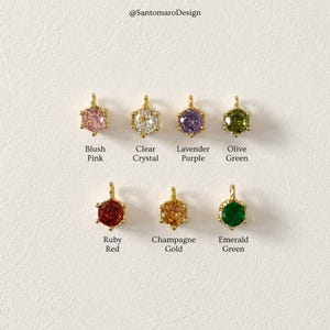 Può includere: Una collezione di otto ciondoli esagonali con pietre preziose in vari colori, tra cui rosa blush, cristallo trasparente e verde smeraldo. Ogni ciondolo ha un'ansa color oro. Il testo sottostante identifica il colore.