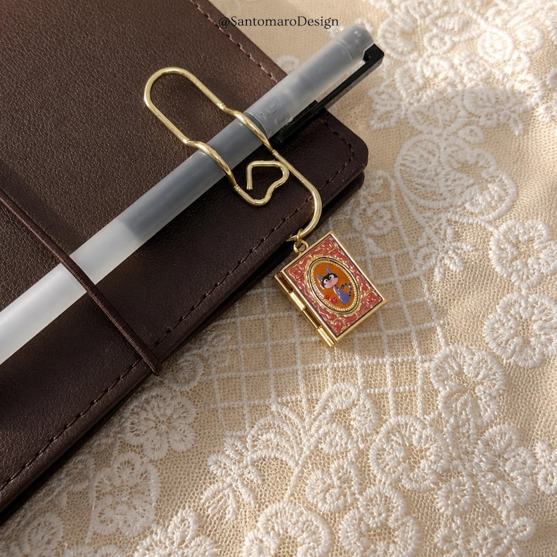 K&ouml;nnte beinhalten: Ein goldfarbener Stiftclip mit herzf&ouml;rmigem Design, der einen Stift und einen kleinen, rechteckigen Anh&auml;nger h&auml;lt. Der Anh&auml;nger zeigt eine farbenfrohe Illustration in einem roten und goldenen Rahmen, befestigt an einem braunen Leder-Notizbuch.