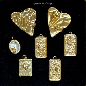 Può includere: Ciondoli dorati in varie forme, tra cui cuori e disegni rettangolari di carte dei tarocchi. Alcuni ciondoli presentano dettagli intricati come un gatto, un sole, una rosa e una mano. I ciondoli sono esposti su una superficie nera.