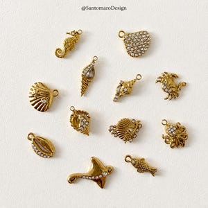 Peut inclure: Divers breloques dorées sur le thème marin, dont un hippocampe, des coquillages et un crabe, sont présentées sur une surface blanche. Certaines breloques sont ornées de cristaux transparents. Les breloques sont conçues avec une boucle pour la joaillerie.