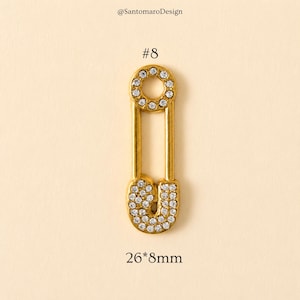 May include: A gold-colored safety pin with a circle and a U-shaped bottom, both encrusted with clear rhinestones. The pin is labeled with the number 8 and the dimensions 26mm x 8mm.