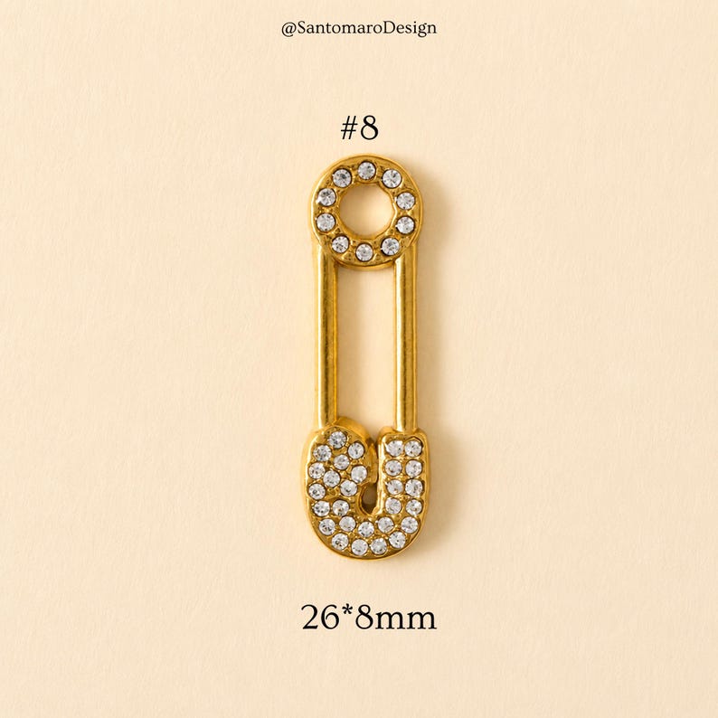 May include: A gold-colored safety pin with a circle and a U-shaped bottom, both encrusted with clear rhinestones. The pin is labeled with the number 8 and the dimensions 26mm x 8mm.