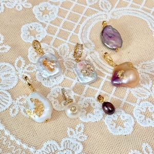 May include: A selection of pearl pendants with gold-coloured detailing. The pearls are in various shapes and colours, including white, pink, purple, and brown. Some pendants have additional embellishments. The image includes the text "@SantomaroDesign".