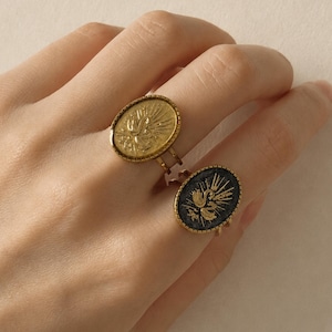 Könnte beinhalten: Zwei ovale Ringe mit goldfarbenen Metallbändern und Fassungen. Ein Ring hat eine goldfarbene Oberfläche mit einem geprägten Schwan- und Sonnenstrahlenmotiv. Der zweite Ring besitzt eine dunkle, texturierte Oberfläche mit einem passenden goldenen Motiv.