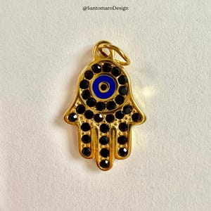 May include: A gold-coloured Hamsa hand charm featuring a blue eye design at its centre. The charm is decorated with black gemstones around the eye and along the fingers. It has a loop at the top for hanging, ideal for jewellery.