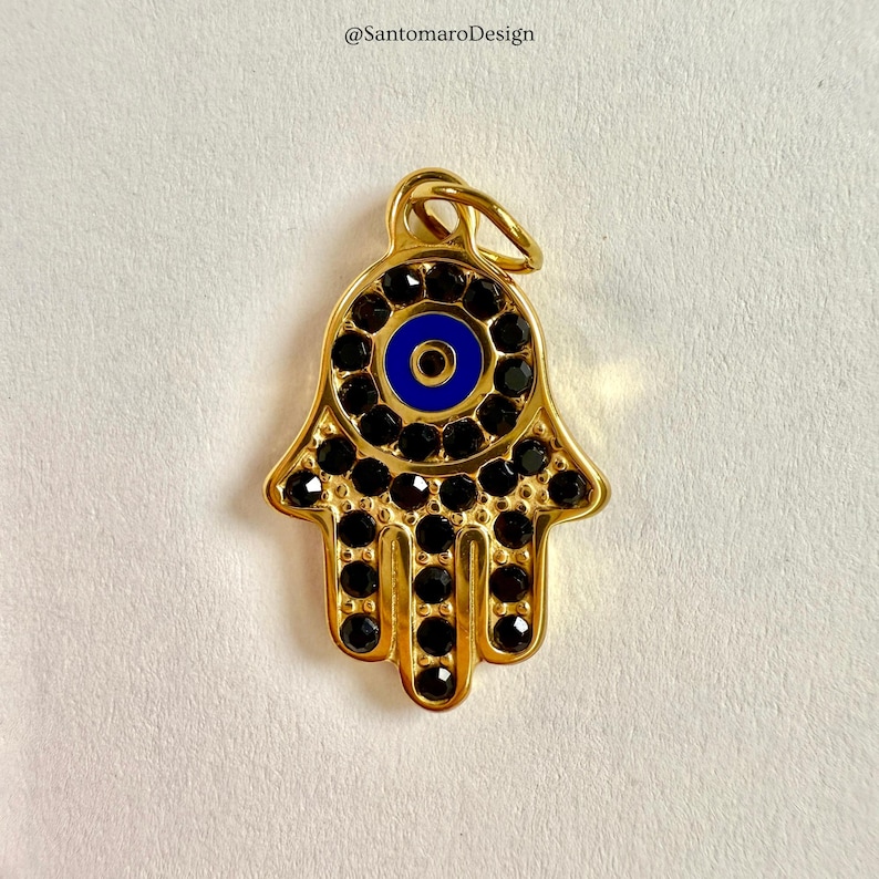 May include: A gold-coloured Hamsa hand charm featuring a blue eye design at its centre. The charm is decorated with black gemstones around the eye and along the fingers. It has a loop at the top for hanging, ideal for jewellery.