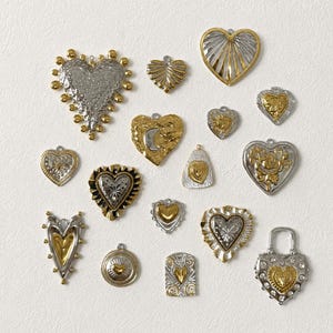 Op de afbeelding: Een verzameling van diverse hartvormige metalen bedels in goud- en zilverkleuren, voorzien van verschillende texturen, reliëfpatronen en decoratieve details. Sommige bedels bevatten kleine heldere steentjes, andere tonen filigraan of hemelse motieven.
