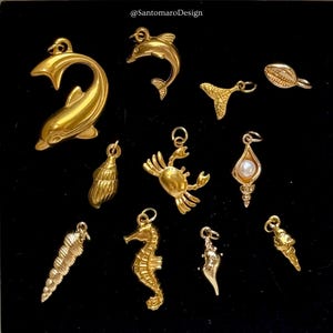 Könnte beinhalten: Eine Sammlung goldfarbener Charms mit Meeresmotiven, darunter Delfine, Muscheln, eine Krabbe, ein Seepferdchen und eine Muschel mit Perle. Die Charms sind vor schwarzem Hintergrund platziert, um ihre detaillierten Designs und ihr glänzendes Finish hervorzuheben.