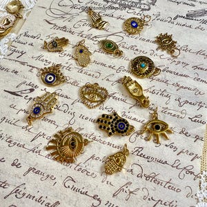 May include: A selection of gold-coloured charms and pendants, including Hamsa hands, evil eyes, and sunburst designs. The charms are embellished with blue, green, and clear gemstones, and are arranged on a vintage paper background with script.