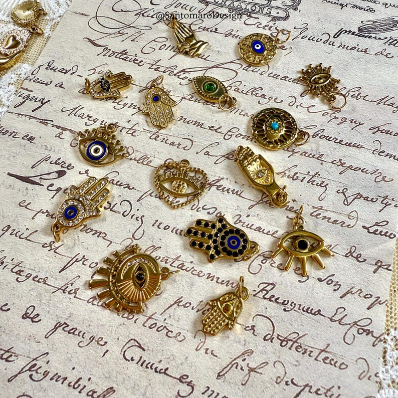 May include: A selection of gold-coloured charms and pendants, including Hamsa hands, evil eyes, and sunburst designs. The charms are embellished with blue, green, and clear gemstones, and are arranged on a vintage paper background with script.