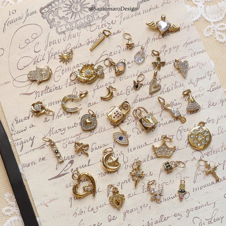 May include: A collection of gold-toned charms with various designs, including hearts, locks, stars, and crowns. Some charms are embellished with clear crystals. The charms are displayed on a vintage paper background.