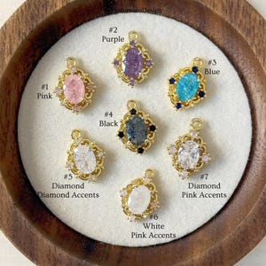 Pode incluir: Uma coleção de sete pingentes dourados ornamentados, cada um adornado com uma pedra preciosa nas cores rosa, roxo, azul, preto e branco. Cada pingente tem uma forma de gema única e é numerado com uma descrição de cor.