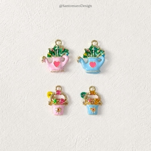 Può includere: Quattro ciondoli smaltati colorati. Due sono annaffiatoi, rosa e blu, con fogliame verde e un cuore. Gli altri due sono cestini di fiori, rosa e blu, con dettagli dorati e fiori colorati. Tutti hanno anelli dorati per appenderli.
