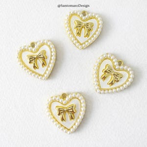 Può includere: Quattro ciondoli a forma di cuore con un centro in smalto bianco e un design a fiocco dorato. Ogni ciondolo è bordato con perle finte e un bordo dorato. I ciondoli hanno un piccolo anello nella parte superiore.