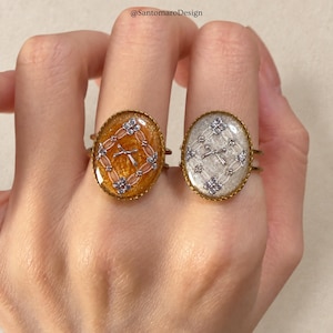 Op de afbeelding: Twee ovale ringen met goudkleurige geschulpte randen. De ene ring heeft een glinsterende oranje harsinleg met een wit ruitpatroon en een centrale strik. De andere ring heeft een glinsterende witte harsinleg met een zilveren ruitpatroon en een centrale strik. De tekst @SantomaroDesign is bovenaan zichtbaar.