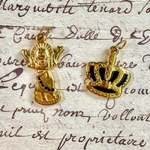 Pu&ograve; includere: Due ciondoli color oro. Uno &egrave; una figura stilizzata con le braccia tese e dettagli neri. L'altro &egrave; una corona con dettagli in smalto nero. Entrambi i ciondoli hanno un anello per una catena.