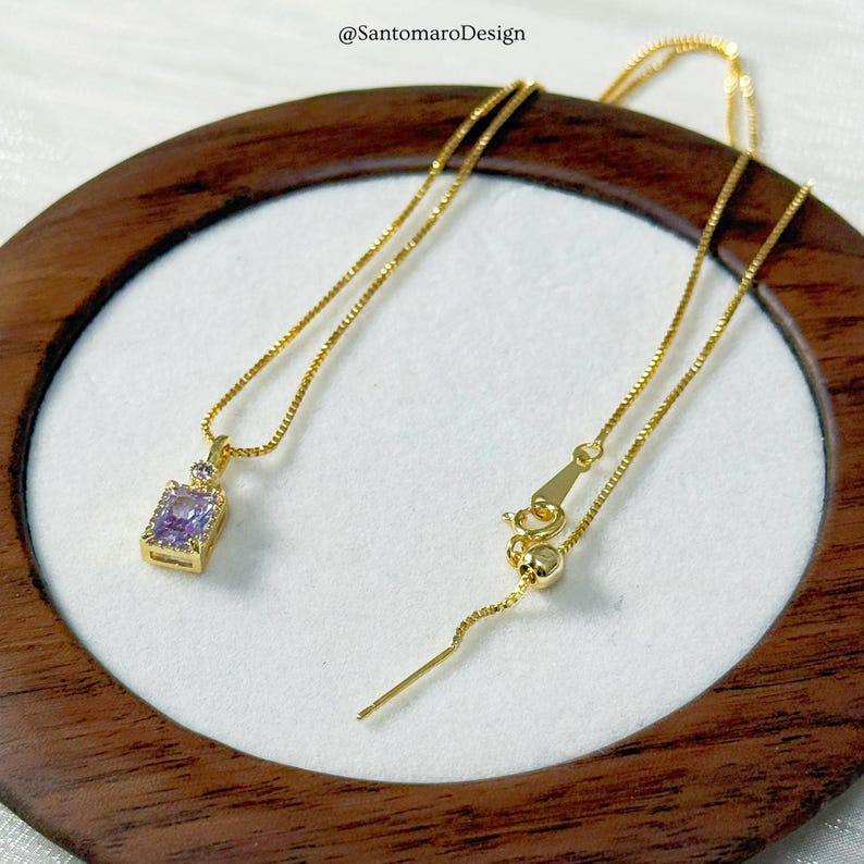 May include: A gold-colored necklace with a rectangular purple gemstone pendant. The pendant is accented with smaller clear stones. The necklace is displayed on a white surface within a wooden frame. The text "@SantomaroDesign" is visible.
