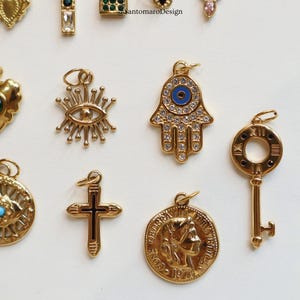 May include: A collection of gold-toned charms, including an eye, a Hamsa hand with a blue eye, a key, a cross, and a coin with a profile. The charms are detailed with gemstones and intricate designs, perfect for jewelry making or adding to existing pieces.