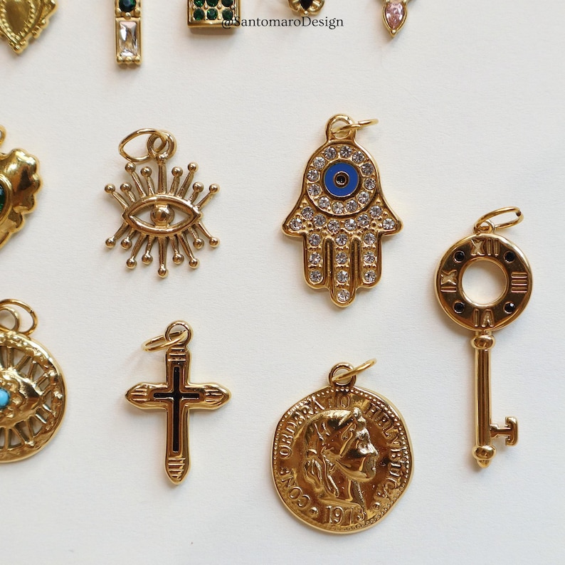 May include: A collection of gold-toned charms, including an eye, a Hamsa hand with a blue eye, a key, a cross, and a coin with a profile. The charms are detailed with gemstones and intricate designs, perfect for jewelry making or adding to existing pieces.