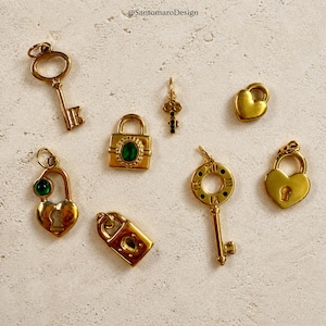 May include: A collection of gold-colored charms, including keys and heart-shaped lockets, are displayed on a textured surface. Some charms feature green gemstone accents. The charms vary in size and design, showcasing intricate details.