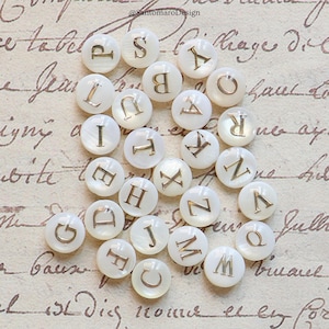 Peut inclure: Un ensemble de boutons ronds blancs, chacun orné d'une lettre dorée de l'alphabet. Les boutons sont disposés sur une surface texturée blanc cassé. L'image comprend le texte "@SantomaroDesign".