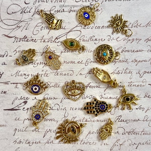 May include: A selection of gold-coloured charms with diverse designs, including hands, eyes, and Hamsa symbols. Some charms feature blue and green accents. The charms are displayed on a vintage paper background.