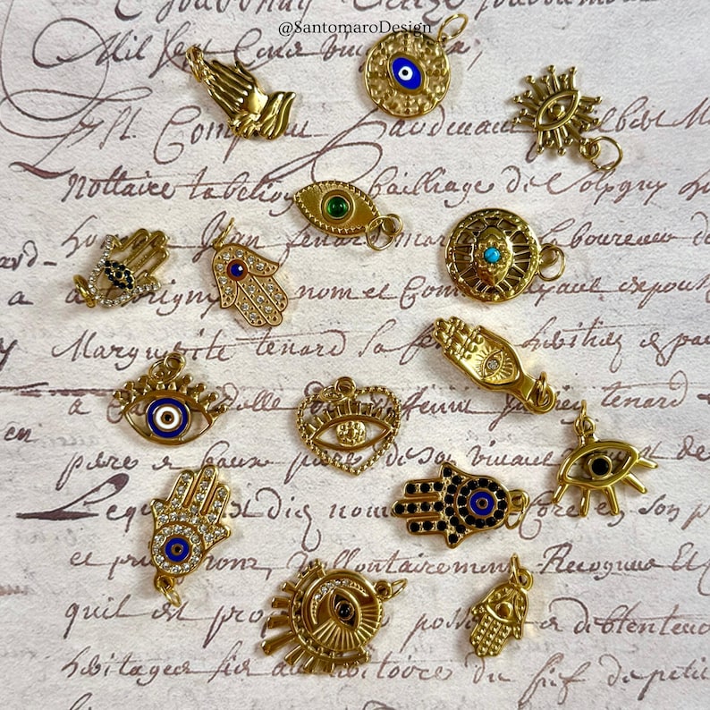 May include: A selection of gold-coloured charms with diverse designs, including hands, eyes, and Hamsa symbols. Some charms feature blue and green accents. The charms are displayed on a vintage paper background.