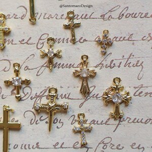 May include: A collection of gold-colored cross charms, some adorned with clear gemstones. The charms vary in design, with intricate details and different shapes. The background features handwritten script and the text "@SantomaroDesign".