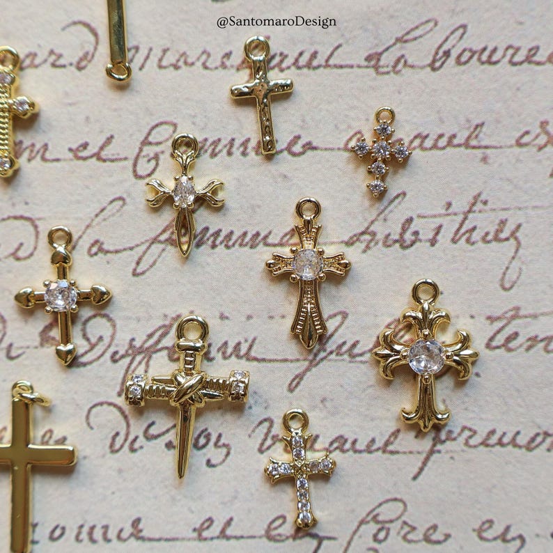 May include: A collection of gold-colored cross charms, some adorned with clear gemstones. The charms vary in design, with intricate details and different shapes. The background features handwritten script and the text "@SantomaroDesign".