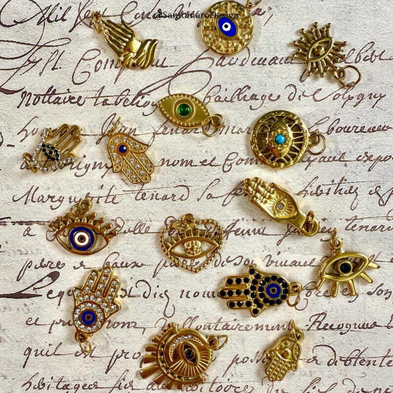 May include: A selection of gold-toned charms and pendants, showcasing diverse designs like hands, eyes, and sunbursts. Some charms have blue, green, and clear crystal embellishments. The charms are presented on a vintage paper backdrop.