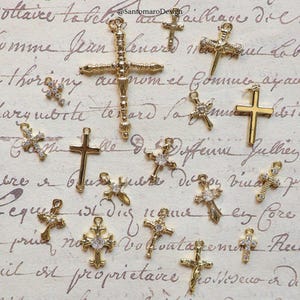 May include: A collection of gold-colored cross charms, some adorned with clear crystals. The crosses vary in design, with some featuring ornate detailing and others a simpler, classic shape. The charms are displayed on a vintage-style paper background.