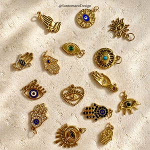 May include: A selection of gold-coloured charms featuring designs such as praying hands, eyes, and Hamsa hands. Some charms have blue and green details, and small gemstones. The charms are arranged on a textured, light-coloured surface.