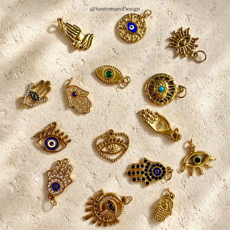 May include: A selection of gold-coloured charms featuring designs such as praying hands, eyes, and Hamsa hands. Some charms have blue and green details, and small gemstones. The charms are arranged on a textured, light-coloured surface.