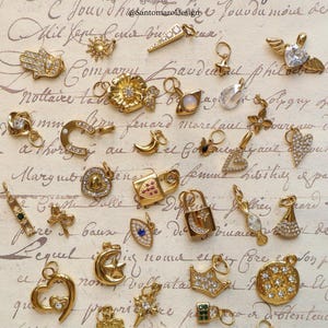 May include: A collection of gold-colored charms and pendants, including a crescent moon with a star, a heart, a lock, and a hand with an eye. The charms are arranged on a vintage paper background with script.