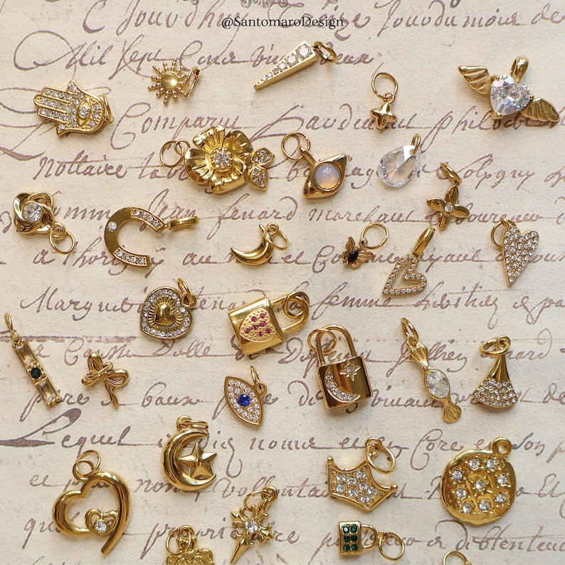 May include: A collection of gold-colored charms and pendants, including a crescent moon with a star, a heart, a lock, and a hand with an eye. The charms are arranged on a vintage paper background with script.
