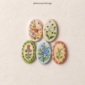 Può includere: Cinque ciondoli ovali in ceramica con disegni floreali dipinti a mano. Ogni pezzo presenta una tavolozza di colori unica che include rosa, bianco, verde, azzurro e crema, con un piccolo foro nella parte superiore. Il testo @SantomaroDesign è visibile.
