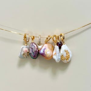 Può includere: Una collezione di pendenti dorati con perle barocche in varie tonalità: bianco, rosa, viola e pesca. Le perle hanno dettagli dorati e sono sospese a una sottile catena d'oro.